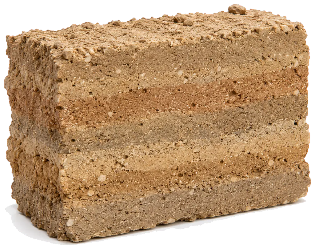 Un bloc de pisé vu de trois quarts, posé sur fond clair. La masse, brun-beige, est divisée en bandes horizontales de teintes légèrement différentes, du beige sable au brun grisé, séparées par des lignes plus claires et plus granuleuses. De petits graviers blancs et ocres affleurent partout. La surface est rugueuse, les arêtes émoussées et irrégulières.
